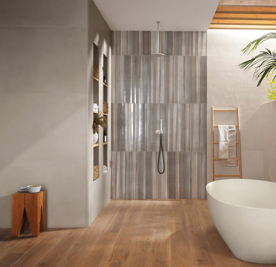 Bathroom with honey wood effect tile floor, beige resin effect wall tiles, shower wall decorated with vertical slabs in shades of gray and beige. white oval basque.